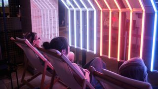 three people sit in chairs, basking in white, orange and blue light coming from three different lamps