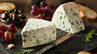 photo shows two large wedges of blue cheese on a cutting board next to a small cheese knife and surrounded by grapes and other fruits