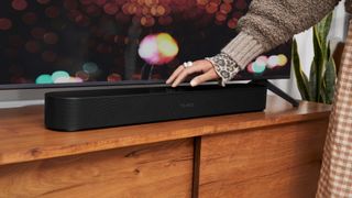 A black Sonos Beam Gen 2 soundbar in front of a TV on a wooden TV cabinet, with a lady resting her hand on it.