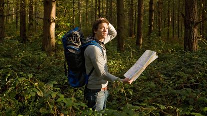 A man hiking in the woods holds a map and looks around, clearly lost.