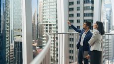 A businessman and businesswoman on the balcony of a skyscraper look at the skyscrapers surrounding them.