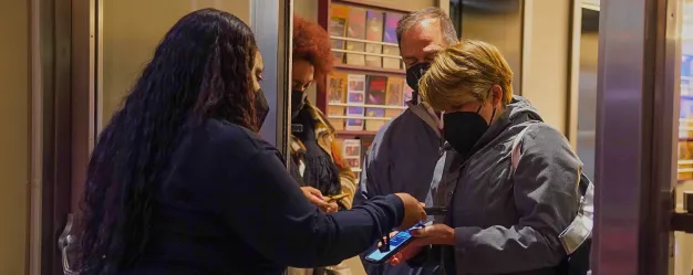 A staff member wearing a black shirt scans tickets for two people dressed in masks and jackets at an entrance.