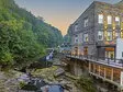 View of the exterior of a hotel that is on the edge of a flowing river. It is made of stone with  a wrap around fence. 