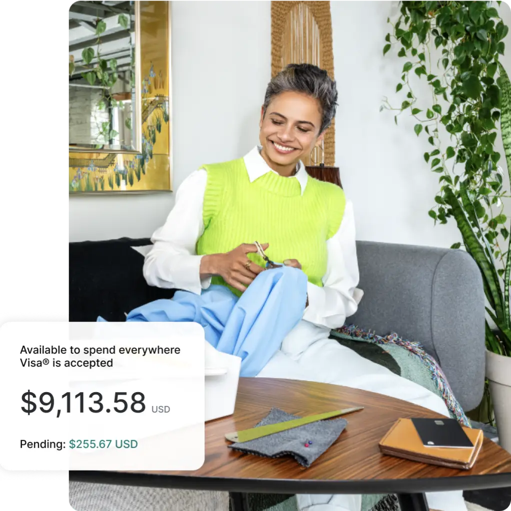 A Spanish woman wearing a bright green vest sits on a sofa smiling while cutting a piece of blue fabric.