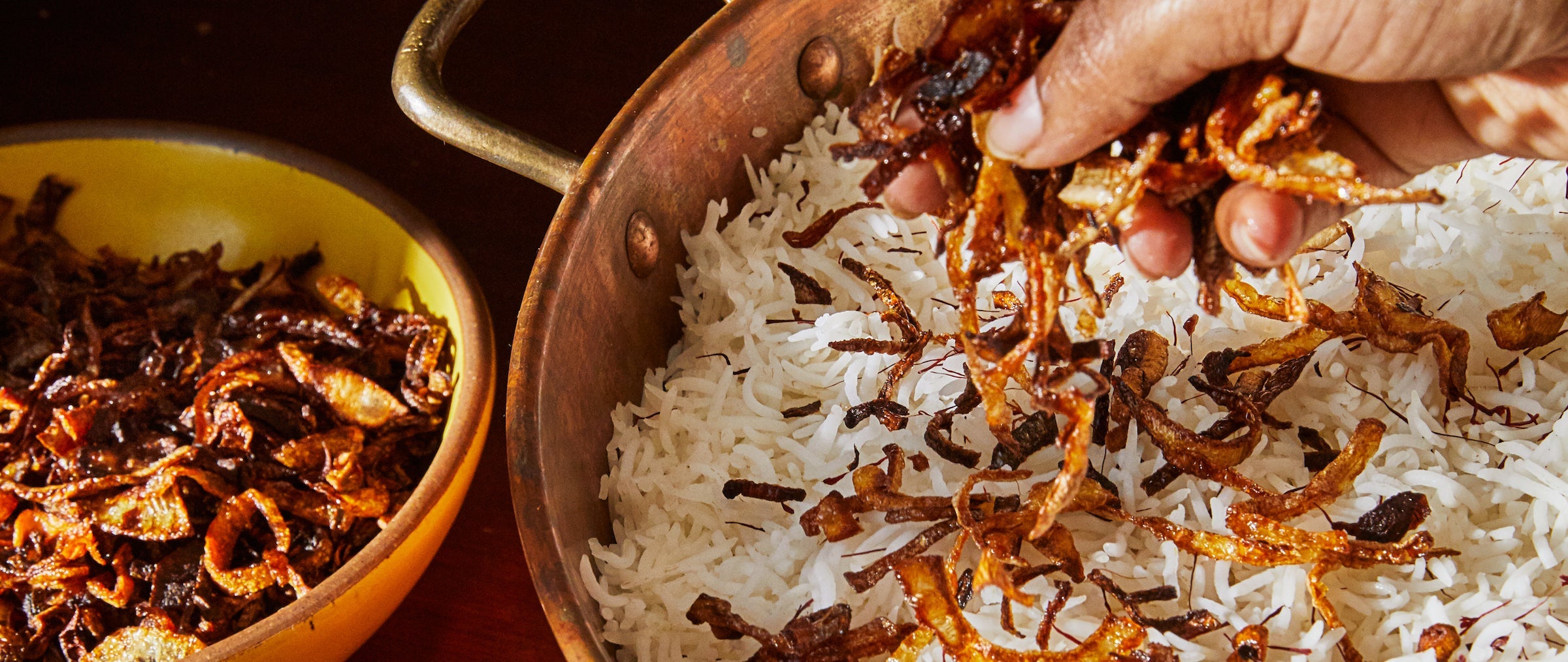 A hand sprinkling fried onions over the rice of a biryani