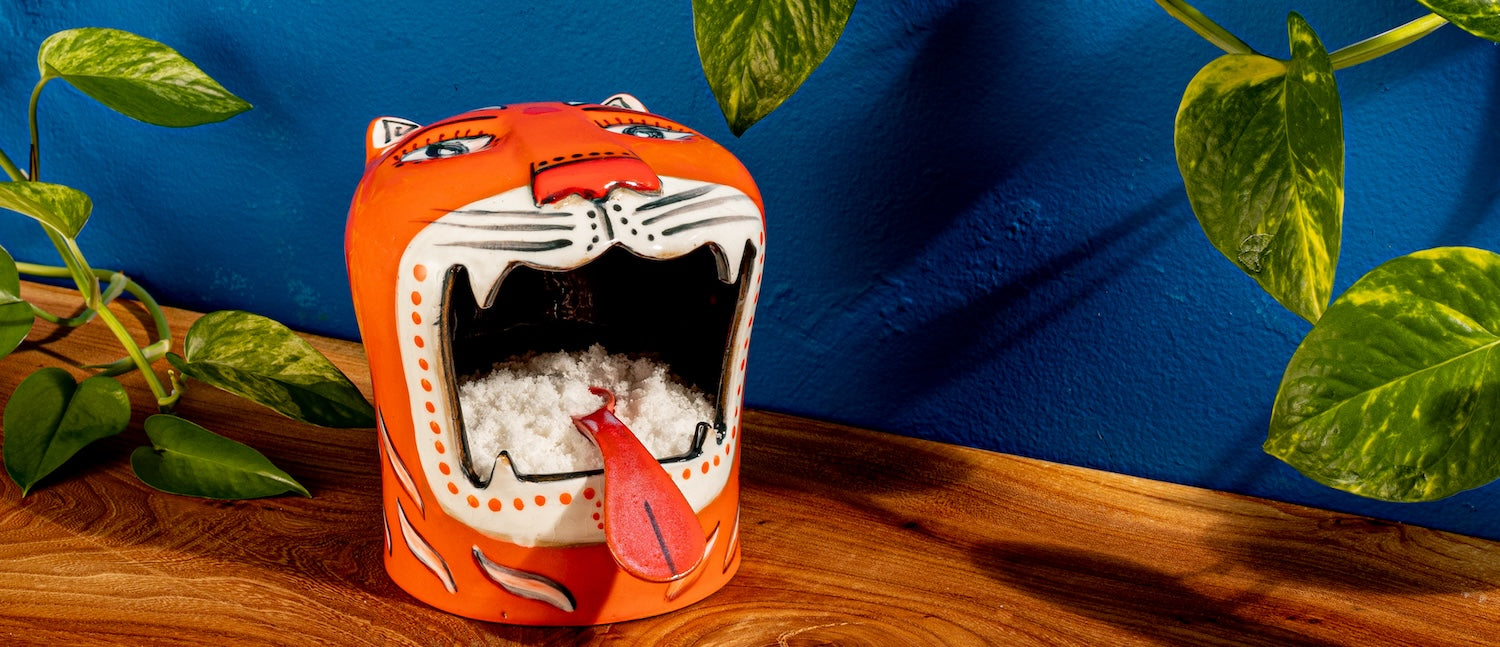 An orange and white ceramic tiger head holds salt in its mouth, on a wood surface with a cobalt blue background and leaves curling around the edges of the frame. 