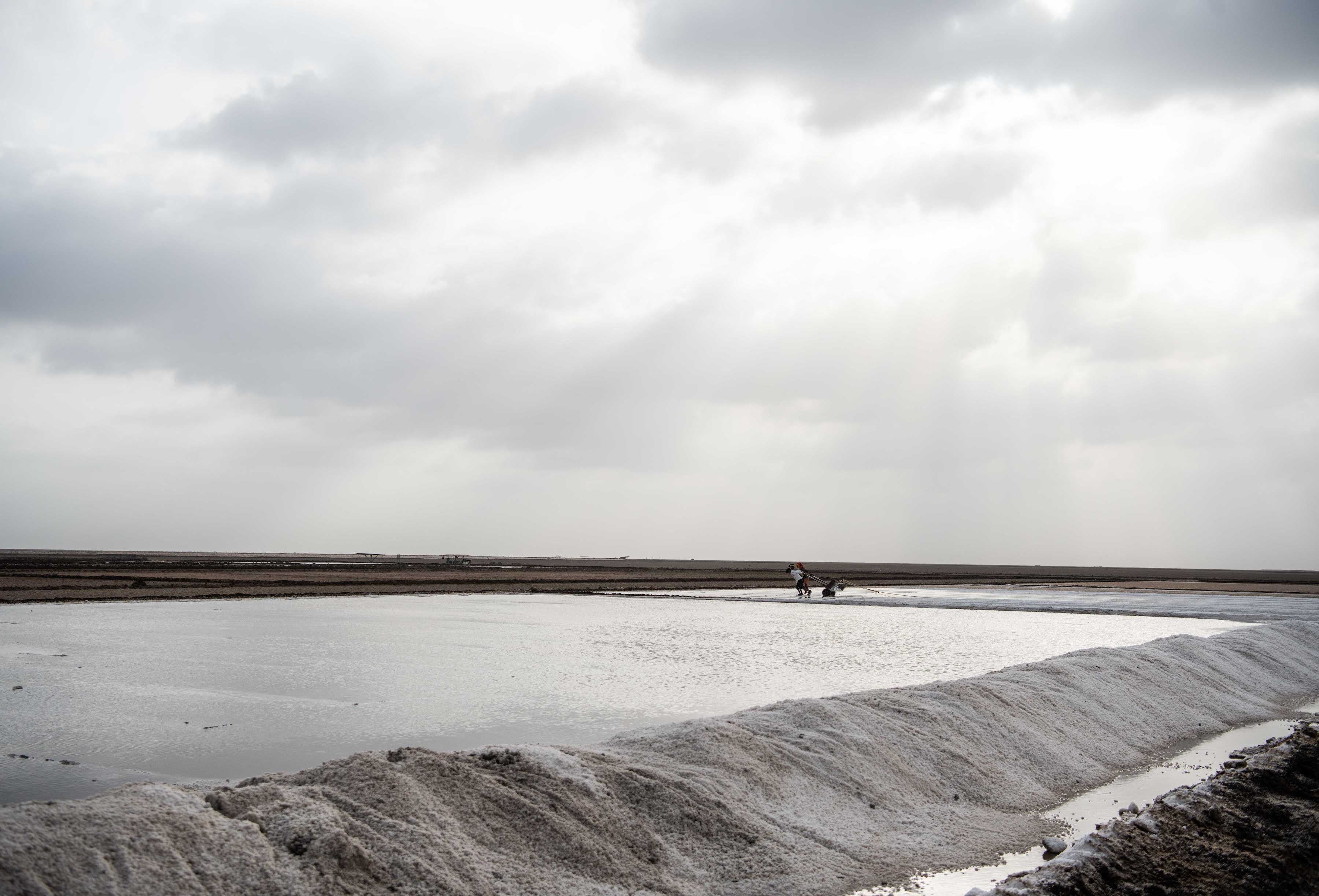 Salt plains - a field of water where far away people are pulling a salt rake on a cloudy day, the light is silvery and streaming from the clouds.