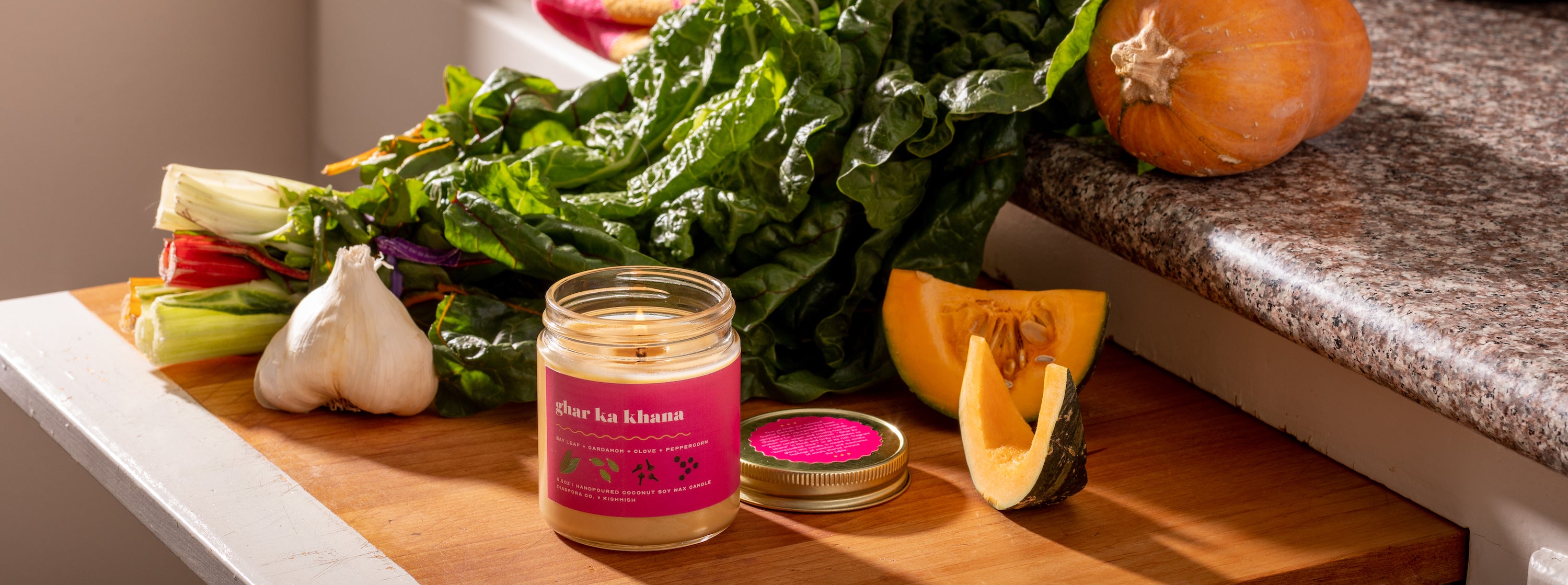 The lit candle on a cutting board with rainbow chard, garlic, and a cut squash in afternoon sunlight