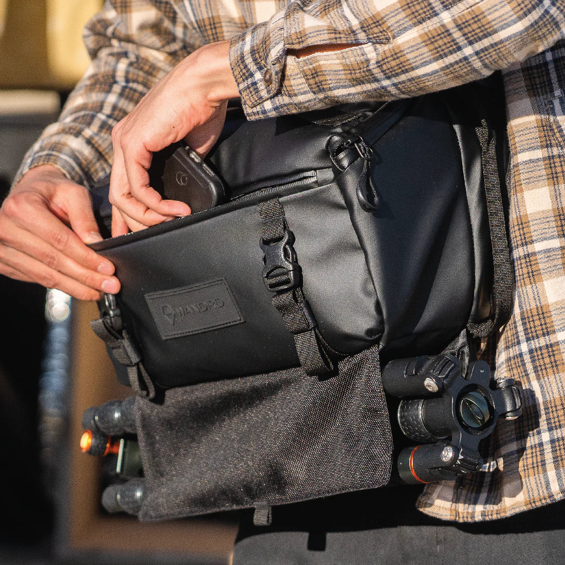 A person wearing the 9 Liter ROAM Sling slides his phone into the front pocket. The bag has a camera tripod attached to the bottom.