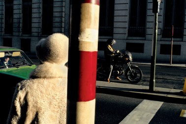 a person in a white fur coat and hat faces away from the camera, toward a green car on the street with a man inside