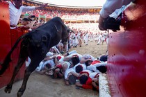 People lie on the ground in a small pile in front of a charging steer that jumps over them into a bullring full of other people.