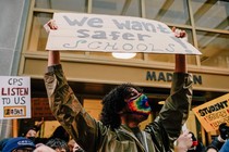 A young man wearing a rainbow mask holds up a sign that says "We want safer schools." In the crowd behind him, other signs read: "CPS Listen to Us" and "Student Voices Matter"