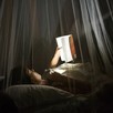 A girl reads a book at night under mosquito netting.