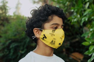 A child with dark curly hair wears a yellow cloth face mask with names of famous scientists and science graphics on it.