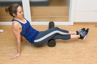 A person in workout clothes rolling the back of their left thigh, with the right leg crossed over top, on a foam roller.