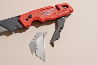 A Milwaukee fastback utility knife with it's blade storage area open. Two spare blades are shown next to the storage area.