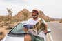 A person reading a road atlas while sitting on the hood of their car, parked on the side of a road in the desert.