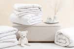 Two white comforters folded up on the floor next to a white teddy bear and one white comforter folded on top of a white ottoman next to an alarm clock and candle on a plate.