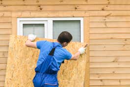 A person wearing gloves cuts a sheet of wood to fit over a set of windows on the exterior of a building.