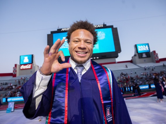 A smiling black man with medium length hair wearing formal clothes and a graduation gown holds up his right hand in the shape of "WC" to represent Wildcats.