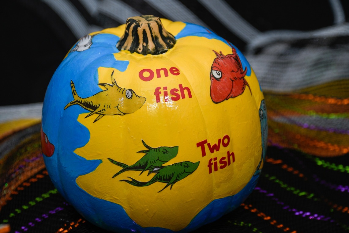 A pumpkin is painted blue and yellow with one yellow fish, one red fish and two bllue fish with the words "one fish" and "two fish" painted on it.