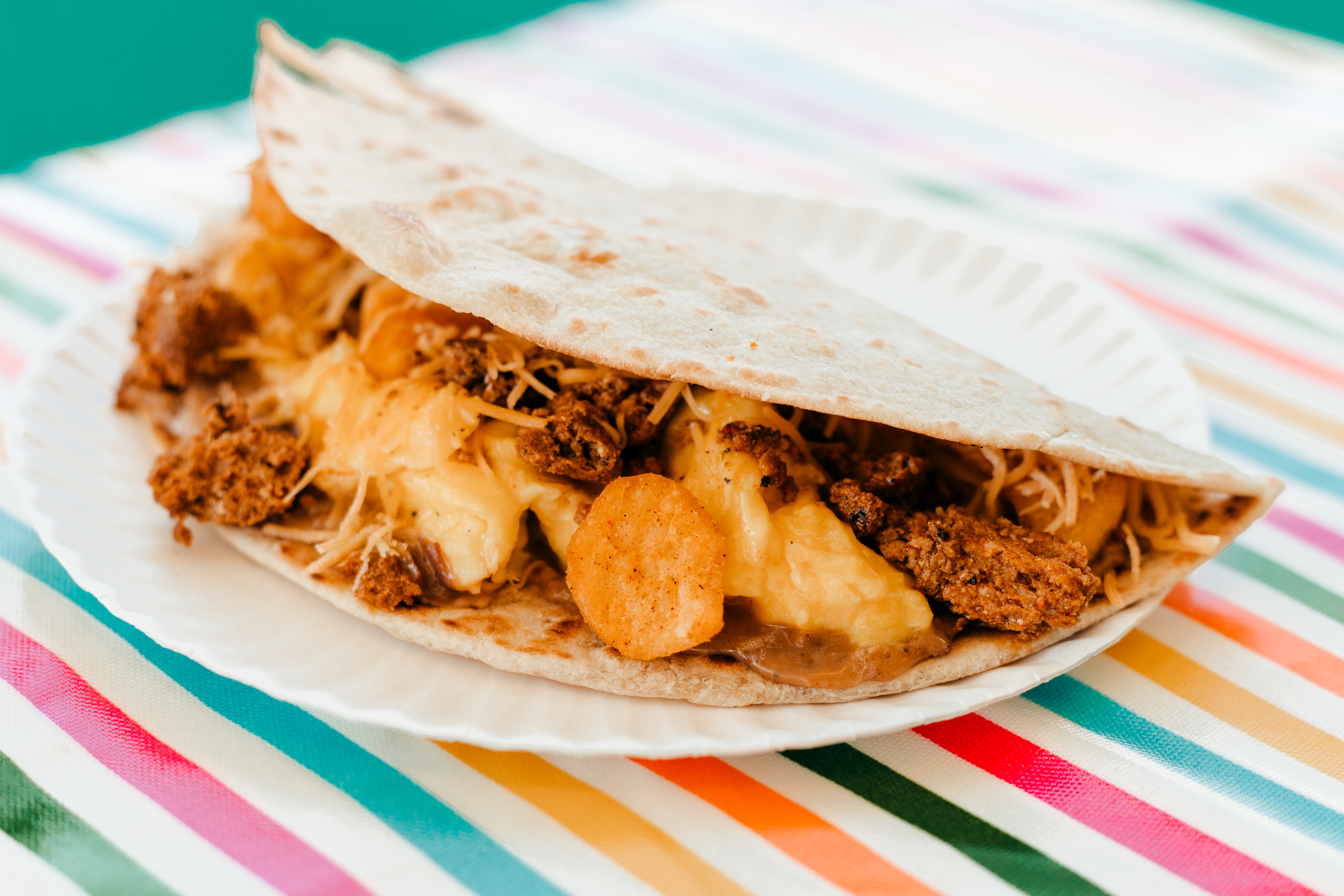 A soft-shell taco stuffed with cheese, ground beef, and potatos sitting on a white paper plate. There’s a pastel-colored stripe tablecloth underneath.