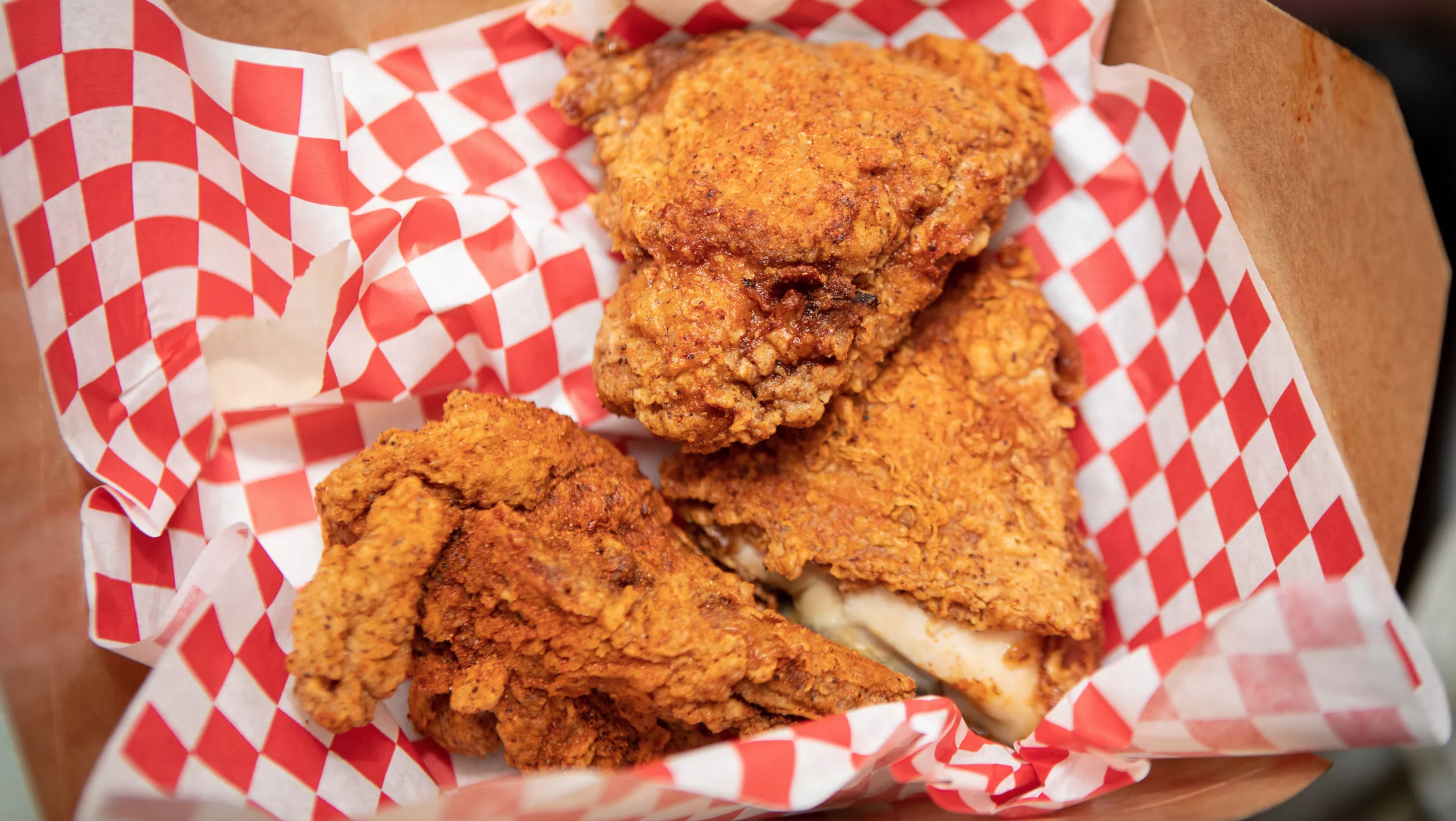 Three pieces of flakey, fried chicken rest in a red-and-white checkered napkin in a takeout basket.