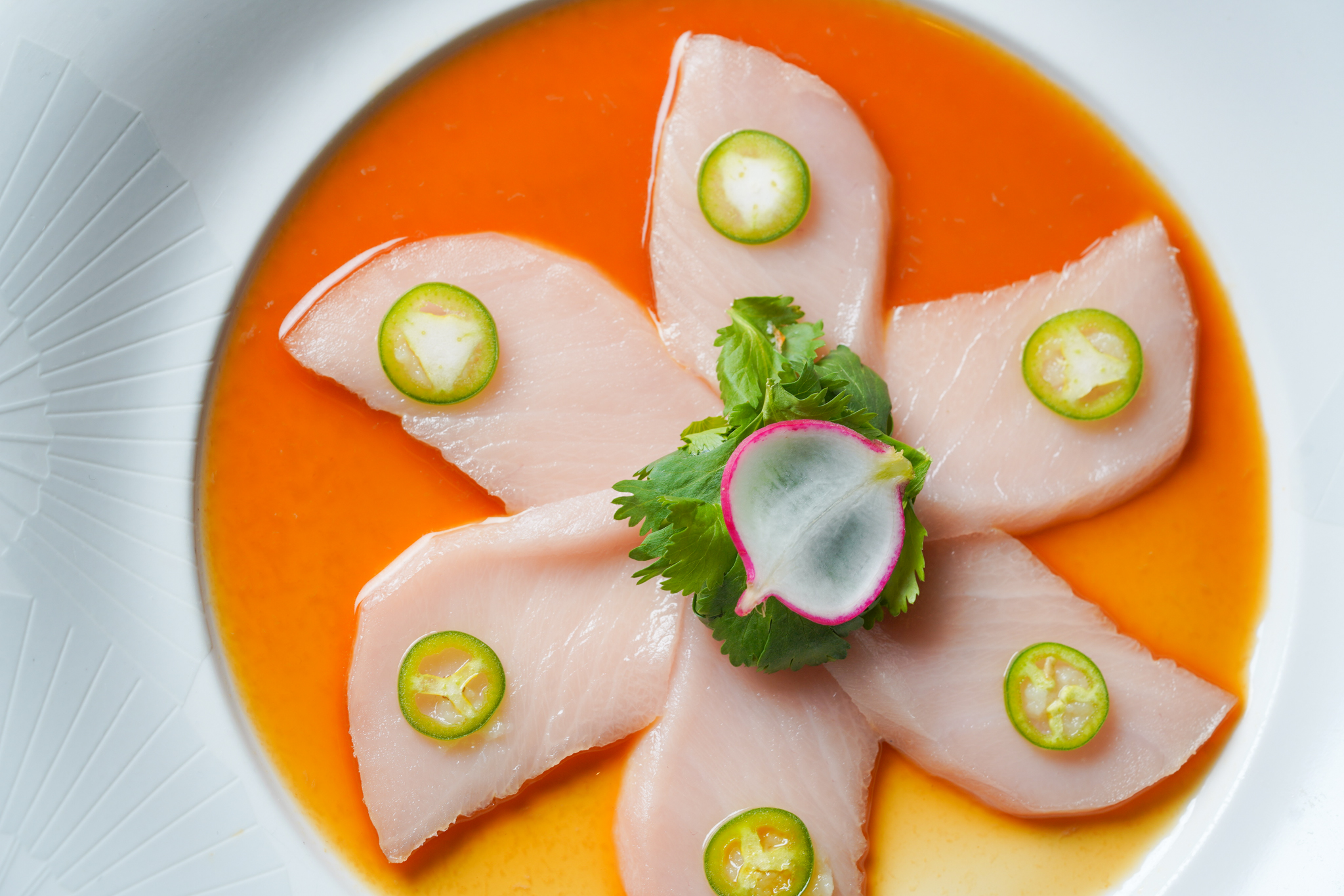 Overhead view of six pieces of raw yellowtail in a pool of light brown sauce, topped with slivers of jalapeño.