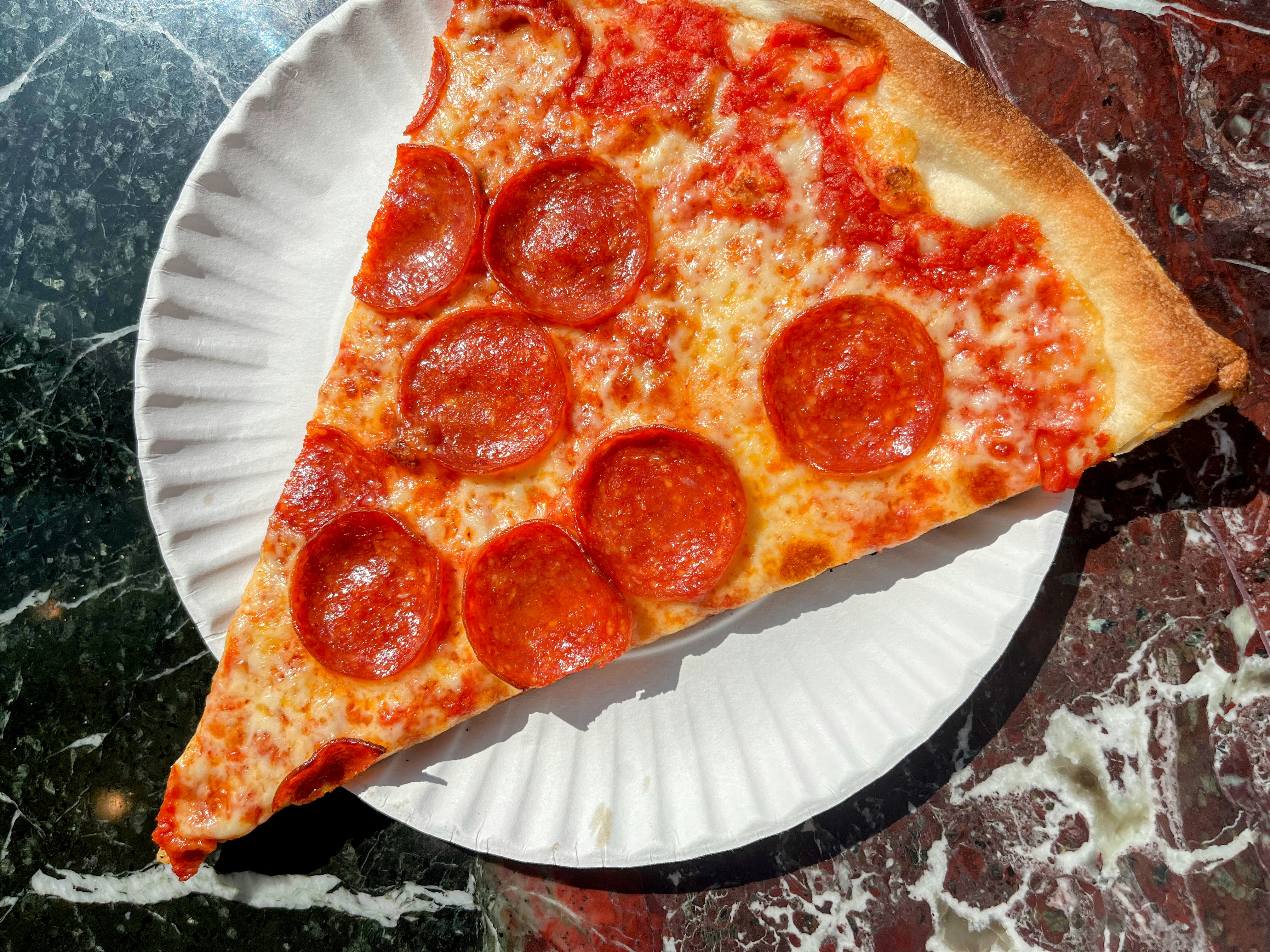 A slice of pepperoni pizza on a white paper plate set on top of a marble counter.