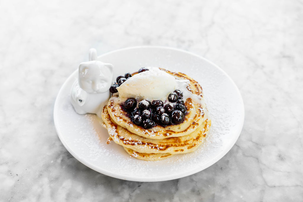 A table of pancakes topped with berries and butter in a lot of syrup next to a cat-shaped pitcher.