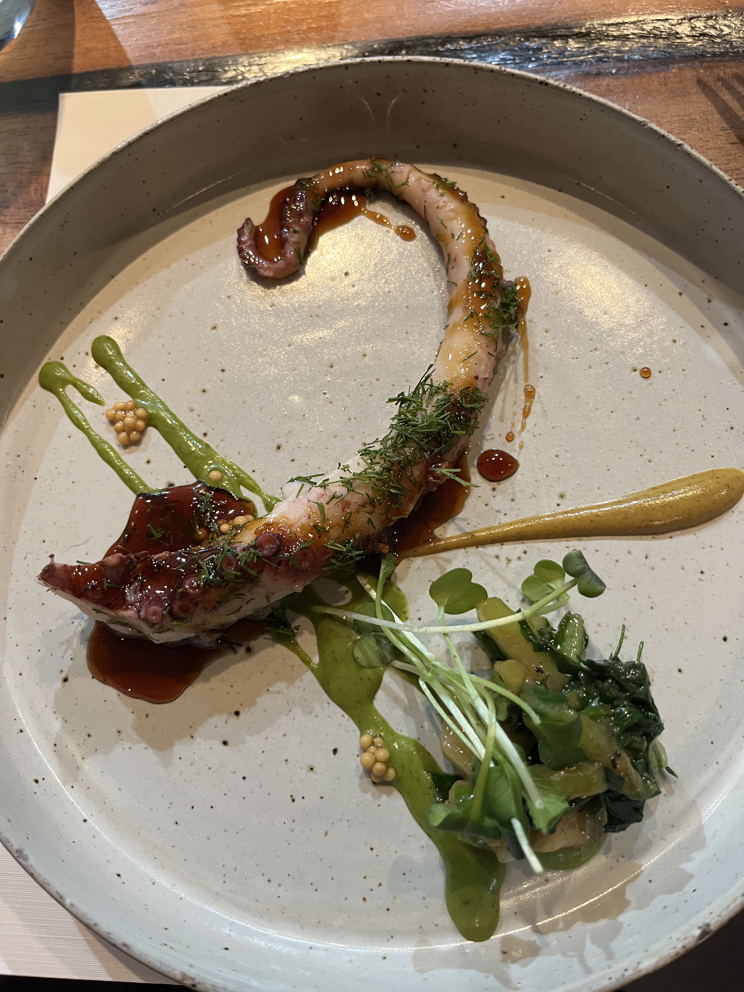 A octopus tentacle on a plate and a side of greens splashed artistically with sauce 