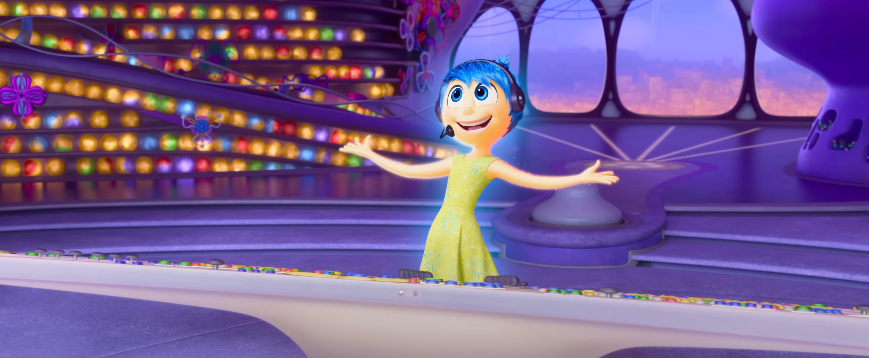 Joy, the yellow-skinned, blue-haired emotion character from the Inside Out movies, stands at the console in emotion HQ, beaming and throwing her arms out in excitement in Inside Out 2
