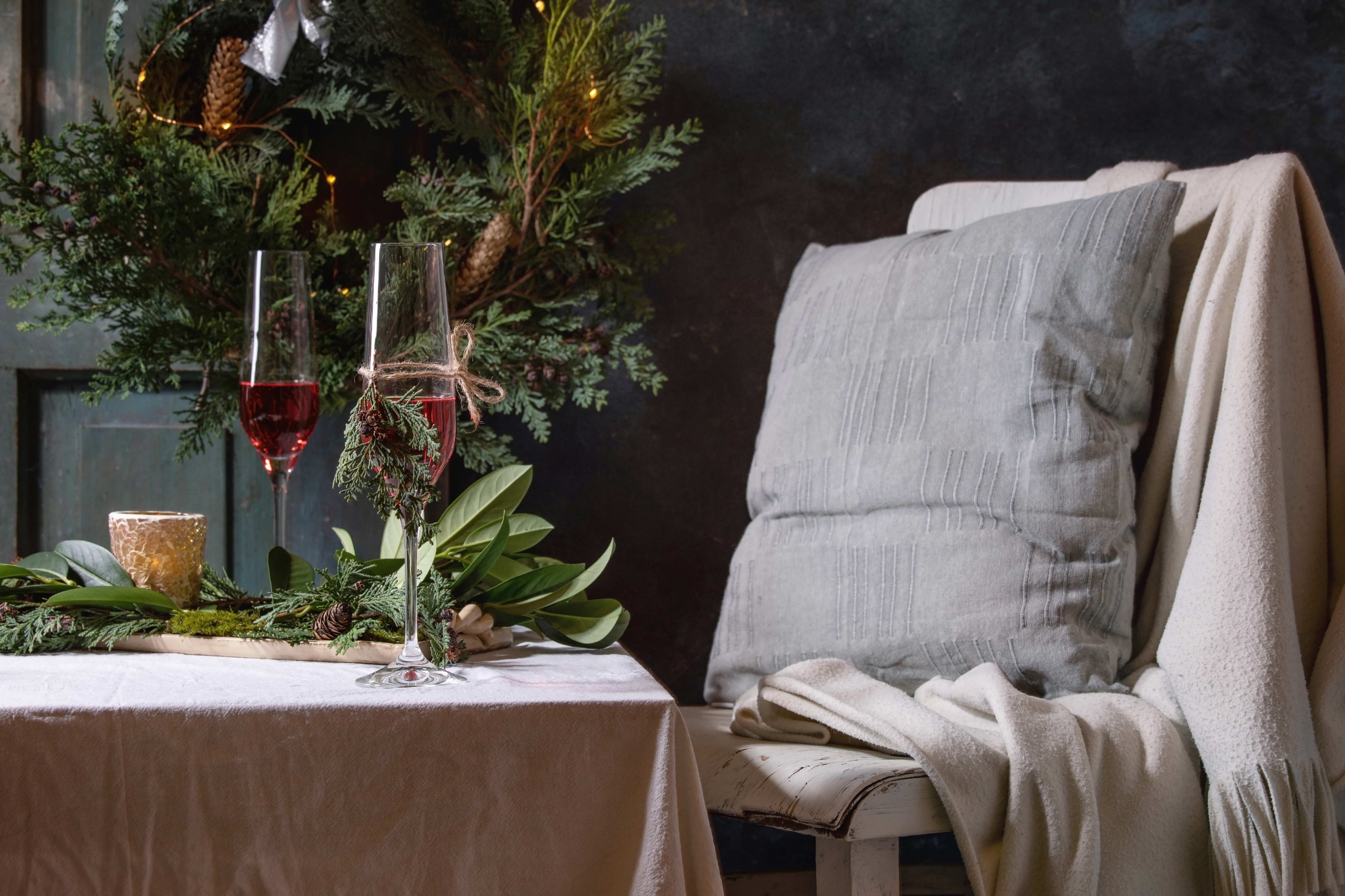 Christmas or New year table setting with two glasses red sparkling wine. plates. napkins. Christmas thuja wreath. luminous garland and burning candles on white tablecloth. Chair with plaid and pillow.