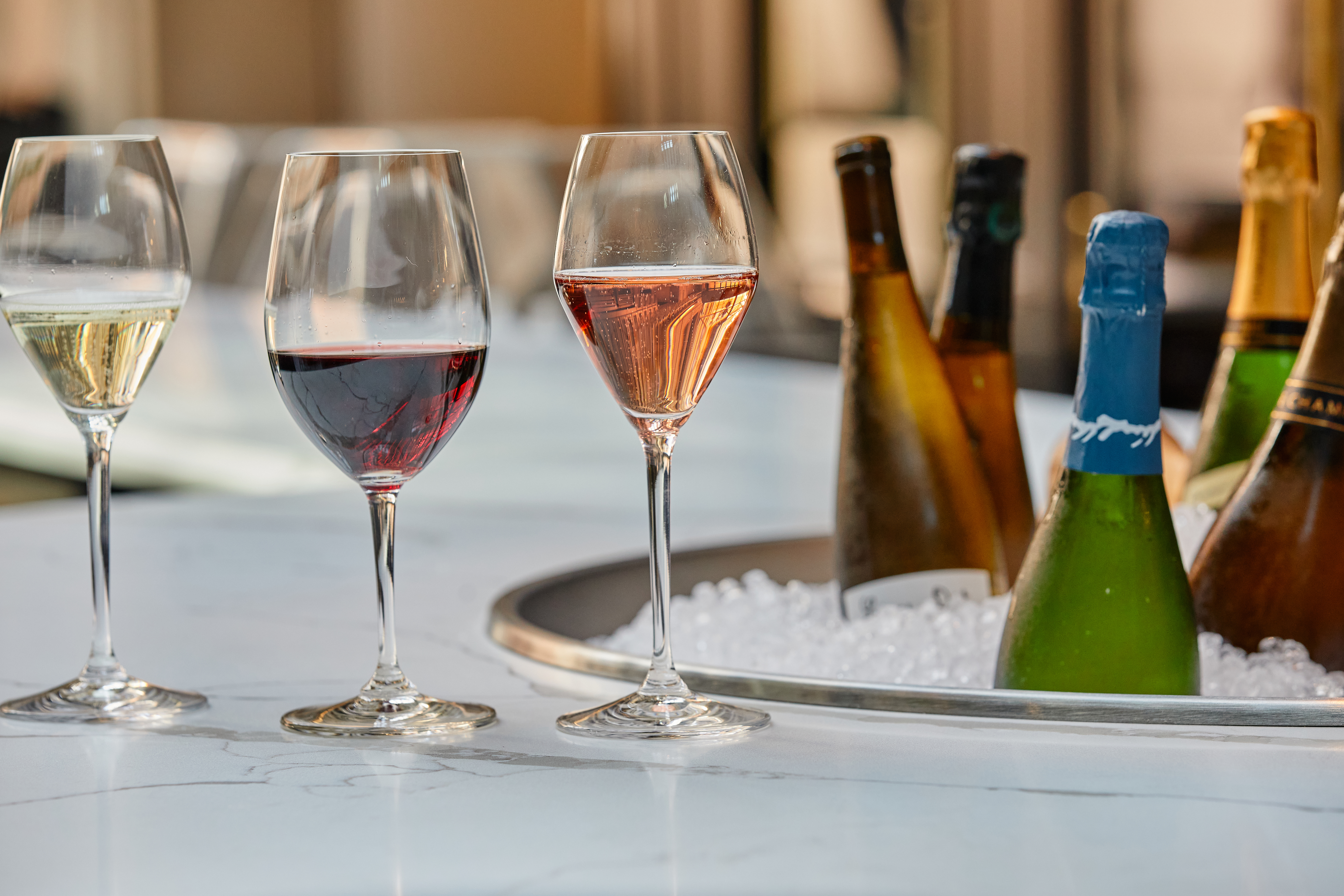 Three glasses of wine (a white, rose, and red) sit on a white bar top next to an ice bath of wine bottles.