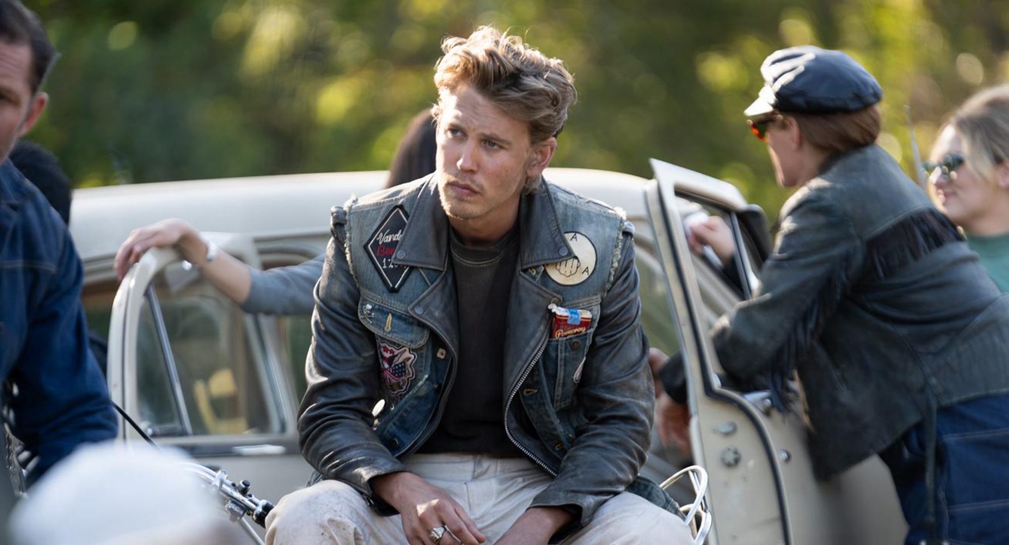 Austin Butler as Benny in The Bikeriders. He sits slouched and scruffy in a jacket covered in patches.