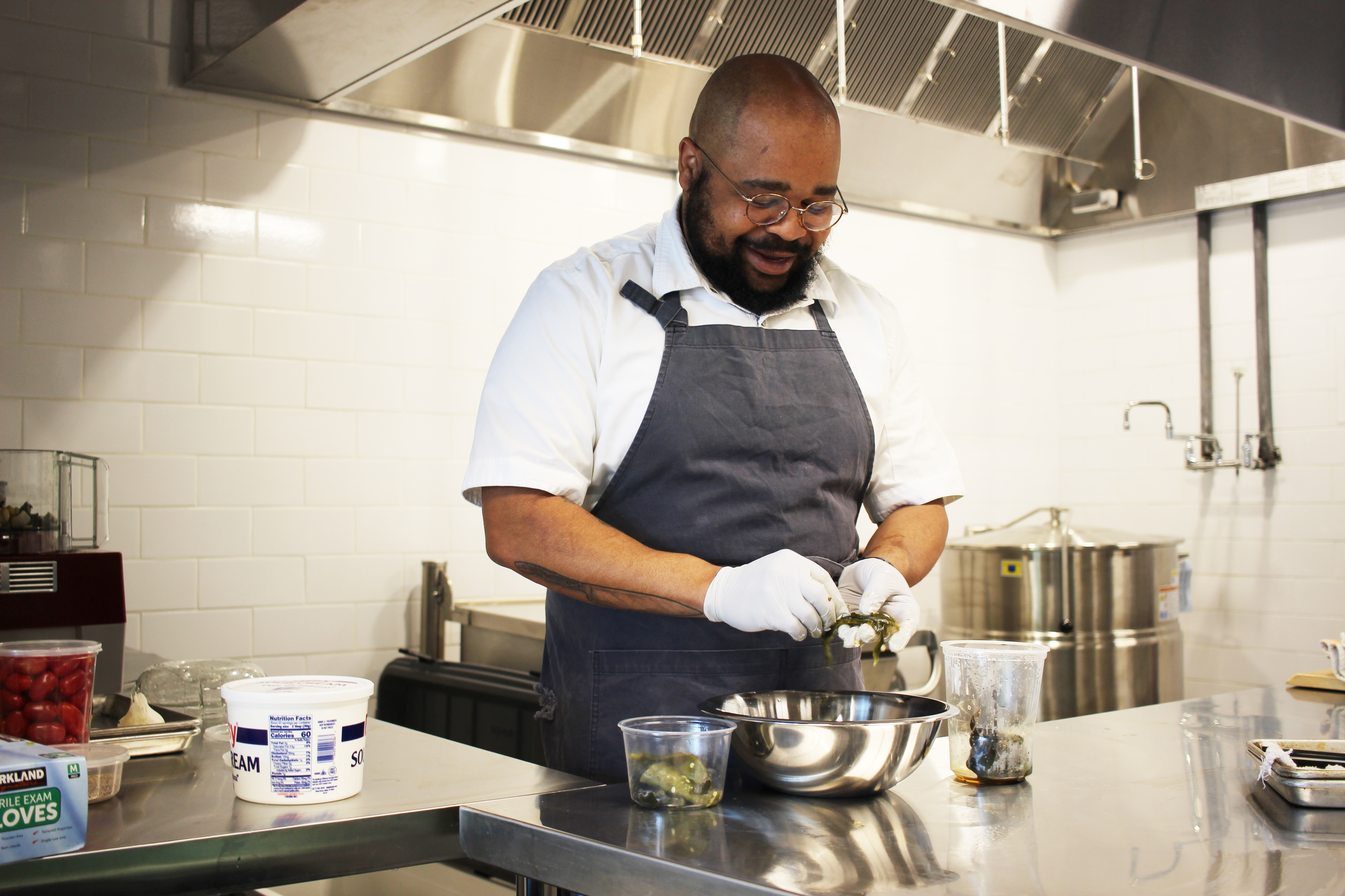 A man wearing an apron working in a kitchen and prepping food to go into a stainless steel bowl sitting on a stainless steel table. 