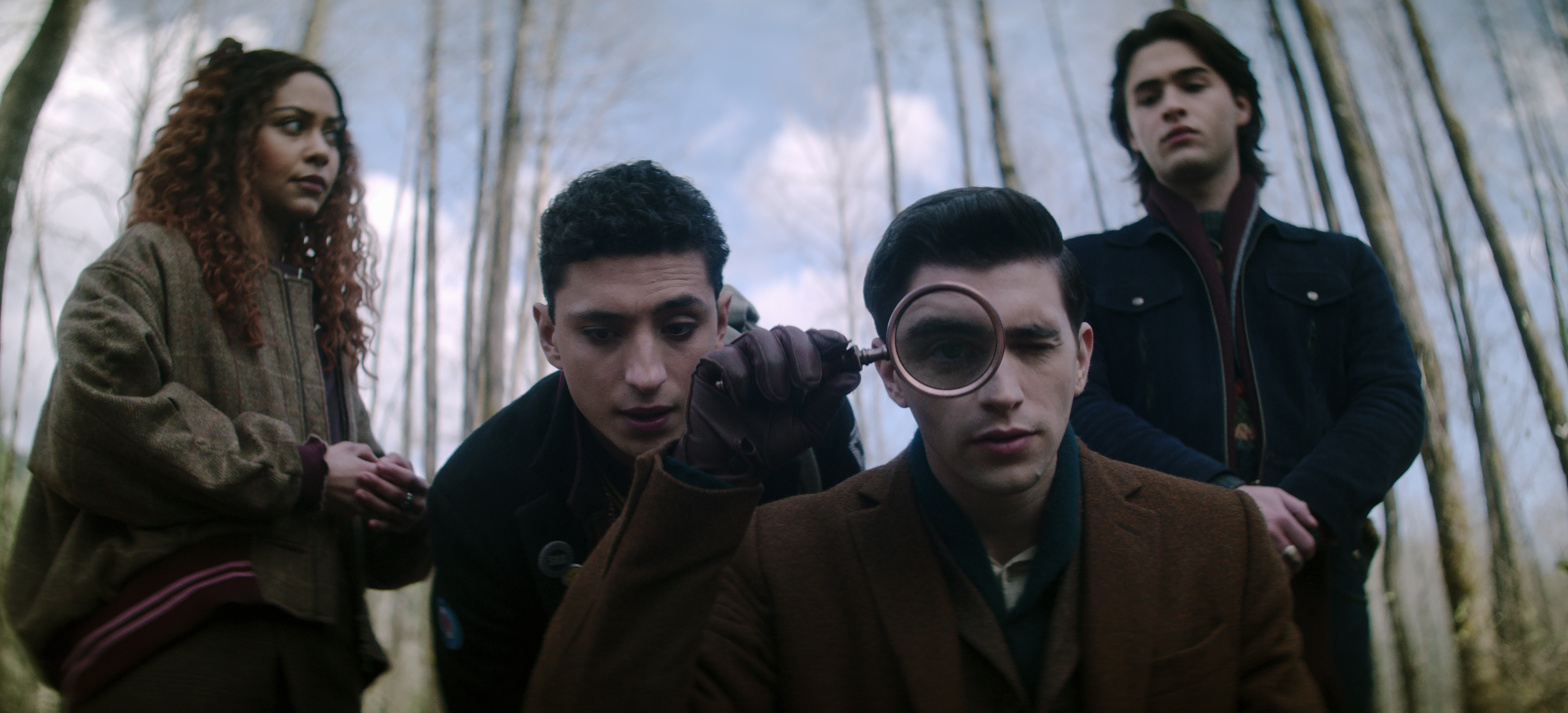 LtR: Kassius Nelson as Crystal Palace, Jayden Revri as Charles Rowland, George Rexstrew as Edwin Payne, and Joshua Colley as Monty in Dead Boy Detectives. Crystal and Monty look skeptical as Charles and Edwin crouch down closer to the camera on the ground. Edwin examines something someone comically with a large magnifying glass held over one eye. 