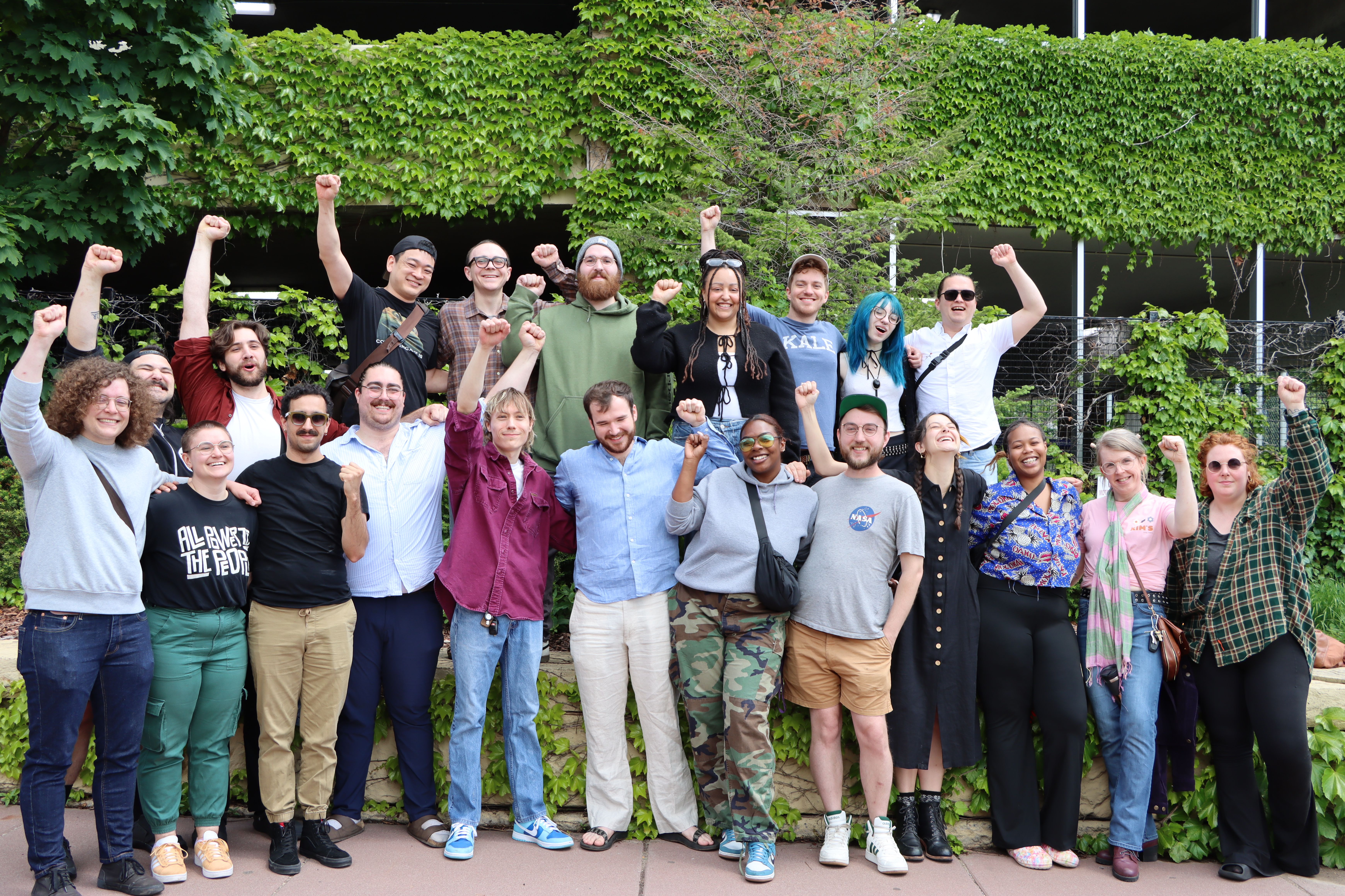 A group of people assembled in front of a parking garage that’s covered with ivy, smiling at the camera with their arms around each other, most of them raising one fist in the air. 