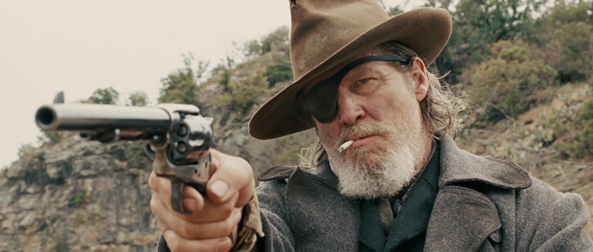 A bearded man with an eyepatch wearing a duster coat and a brown hat holding a revolver in True Grit (2010).