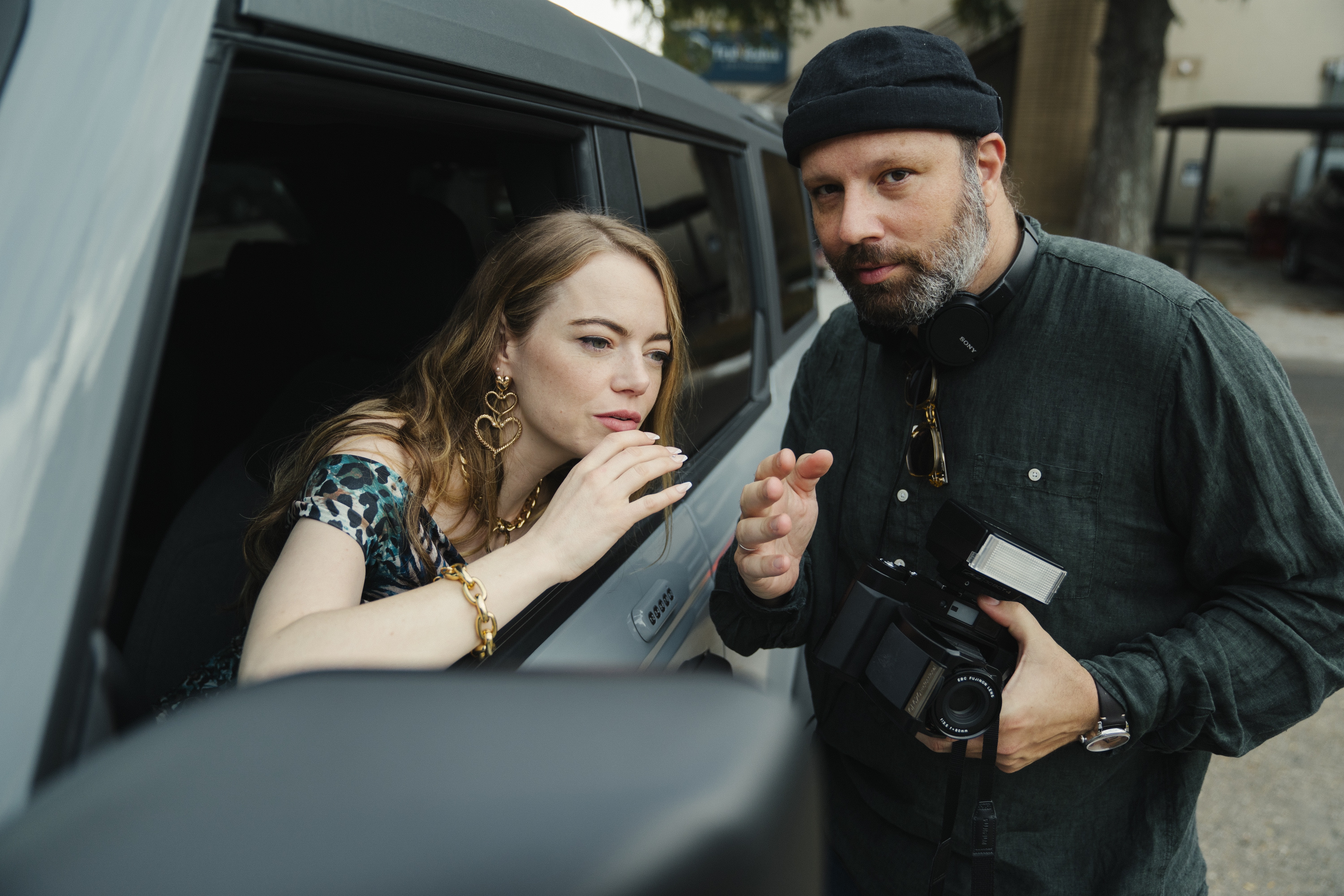 Emma Stone leans out of a car window and talks to director Yorgos Lanthimos, wearing a beanie and carrying a camera, on the set of Kinds of Kindness