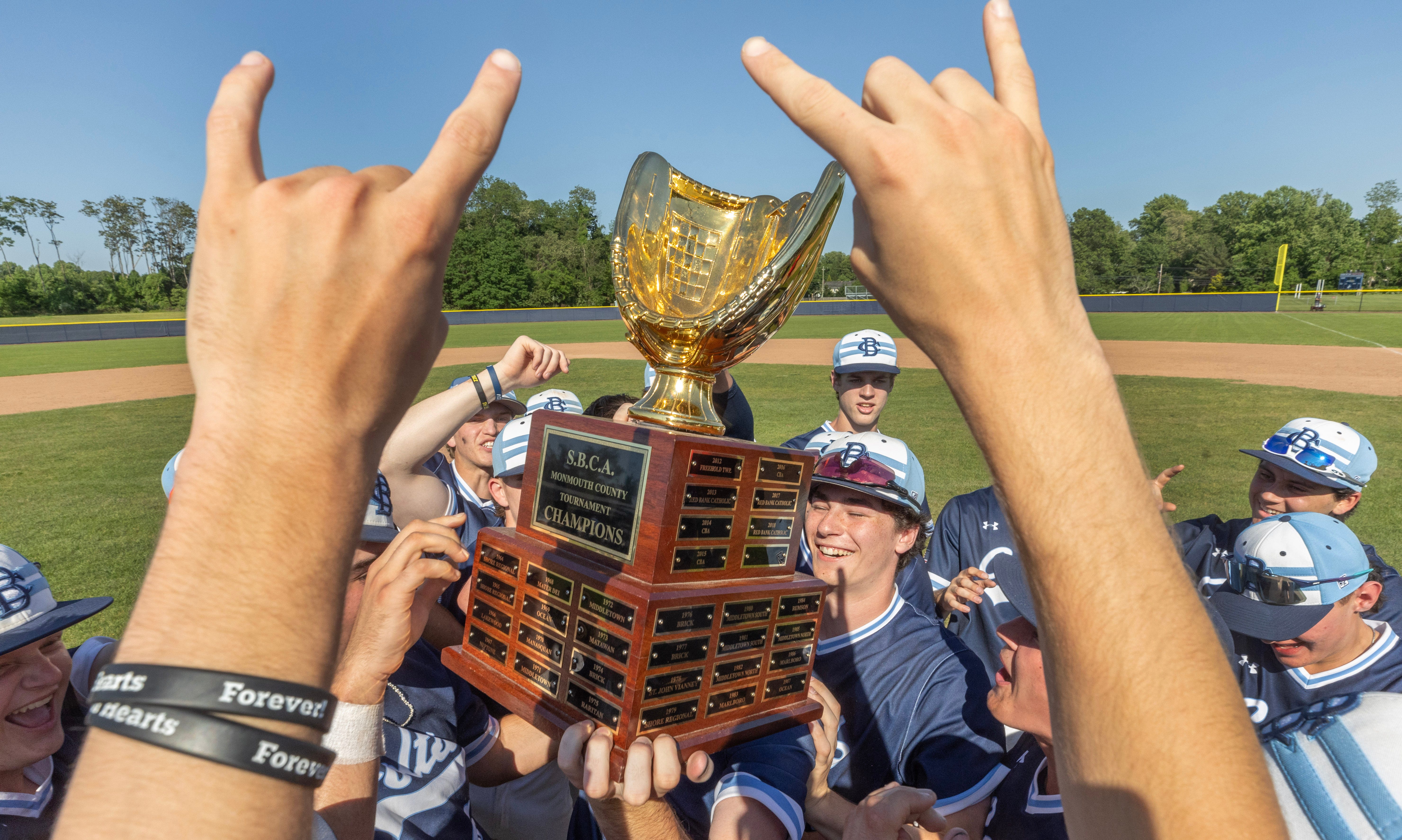 In the foreground, an unseen person throws up the double “devil’s horns” sign, while inbetween the hands, you see a baseball player surrounded by his team and holding a trophy shaped like a baseball glove