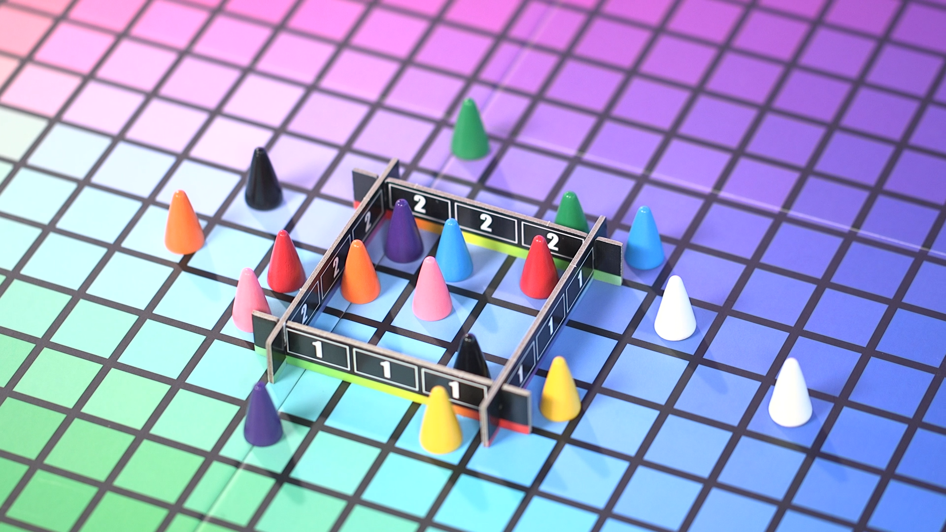 Colorful cone shaped board game pieces sit on grid with a colorful gradient of greens, blues, purples, and pinks. A cardboard square with numbers on it encompasses nine squares on the grid, with some cones inside it and some outside.