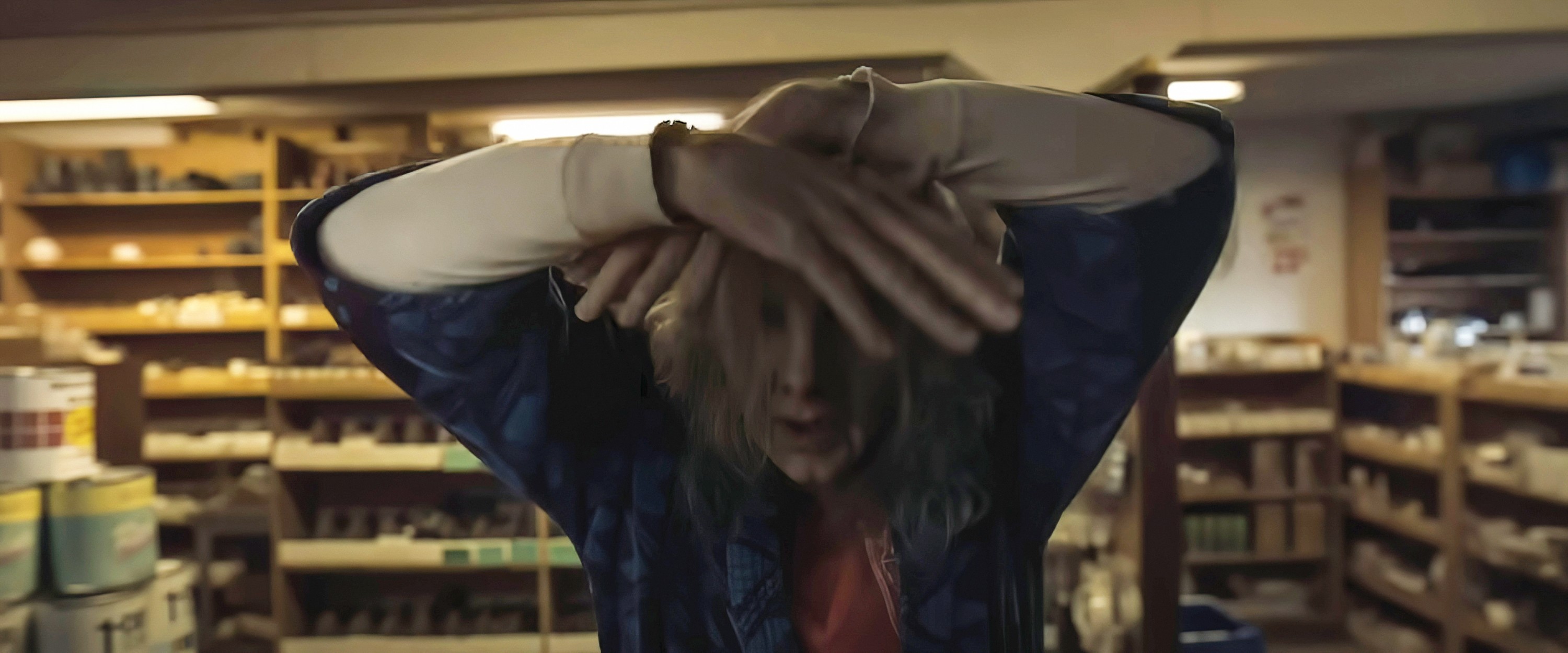 Satanic serial killer Longlegs, a chalk-skinned man with distended, puffy features and long, lank white hair, stands at a hardware store counter, partially covering his face with his hands, in Oz Perkins’ Longlegs