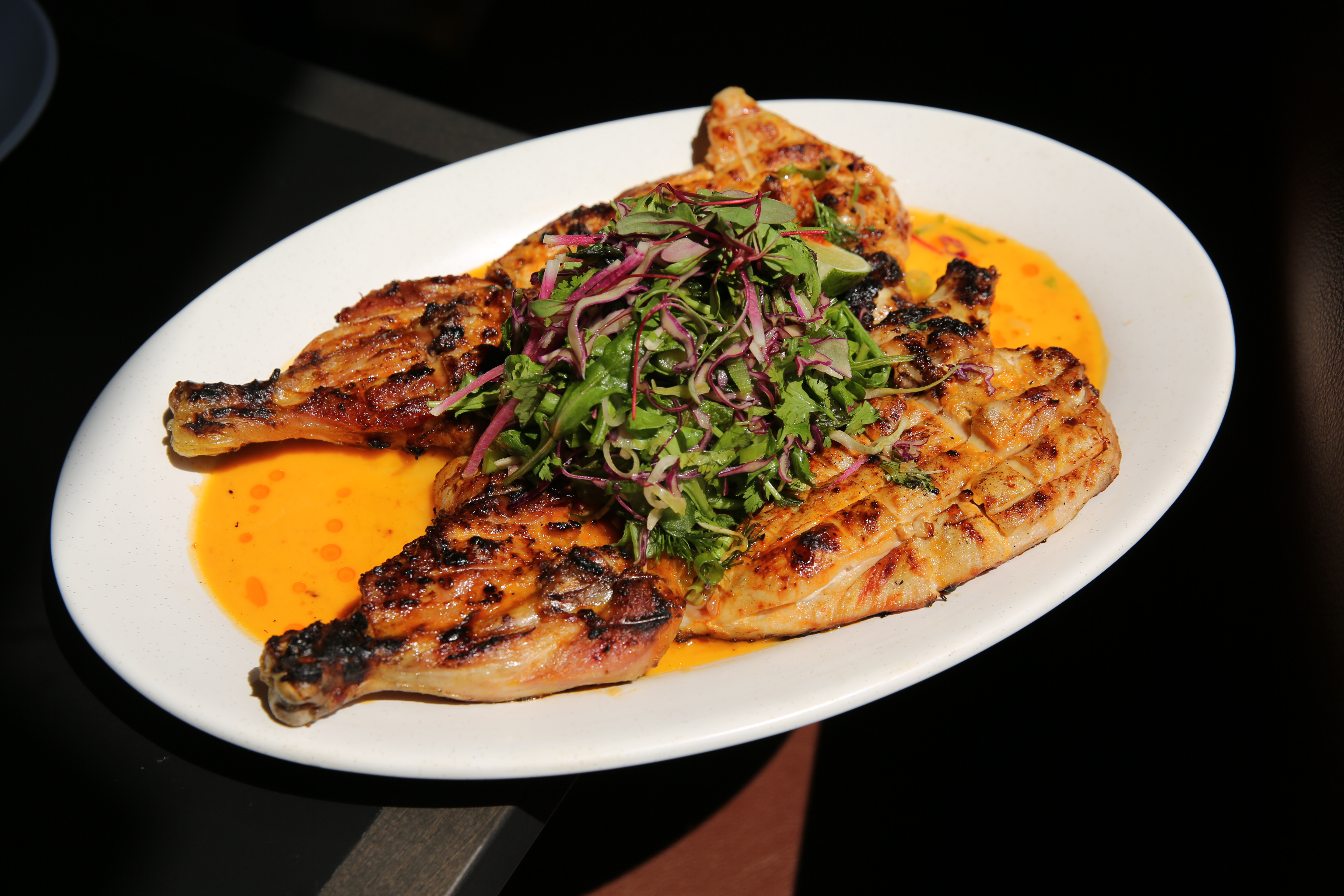 An oval white plate with a whole deboned grilled chicken laid on it flat, sitting in a pool of bright orange sauce with an herb salad on top.