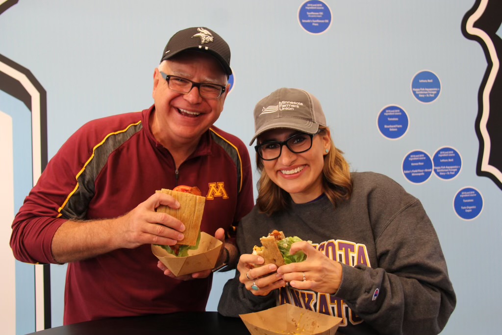 Governor Tim Walz and Lt. Governor Peggy Flanagan smiling, wearing baseball caps, and eating sandwiches against a blue background.