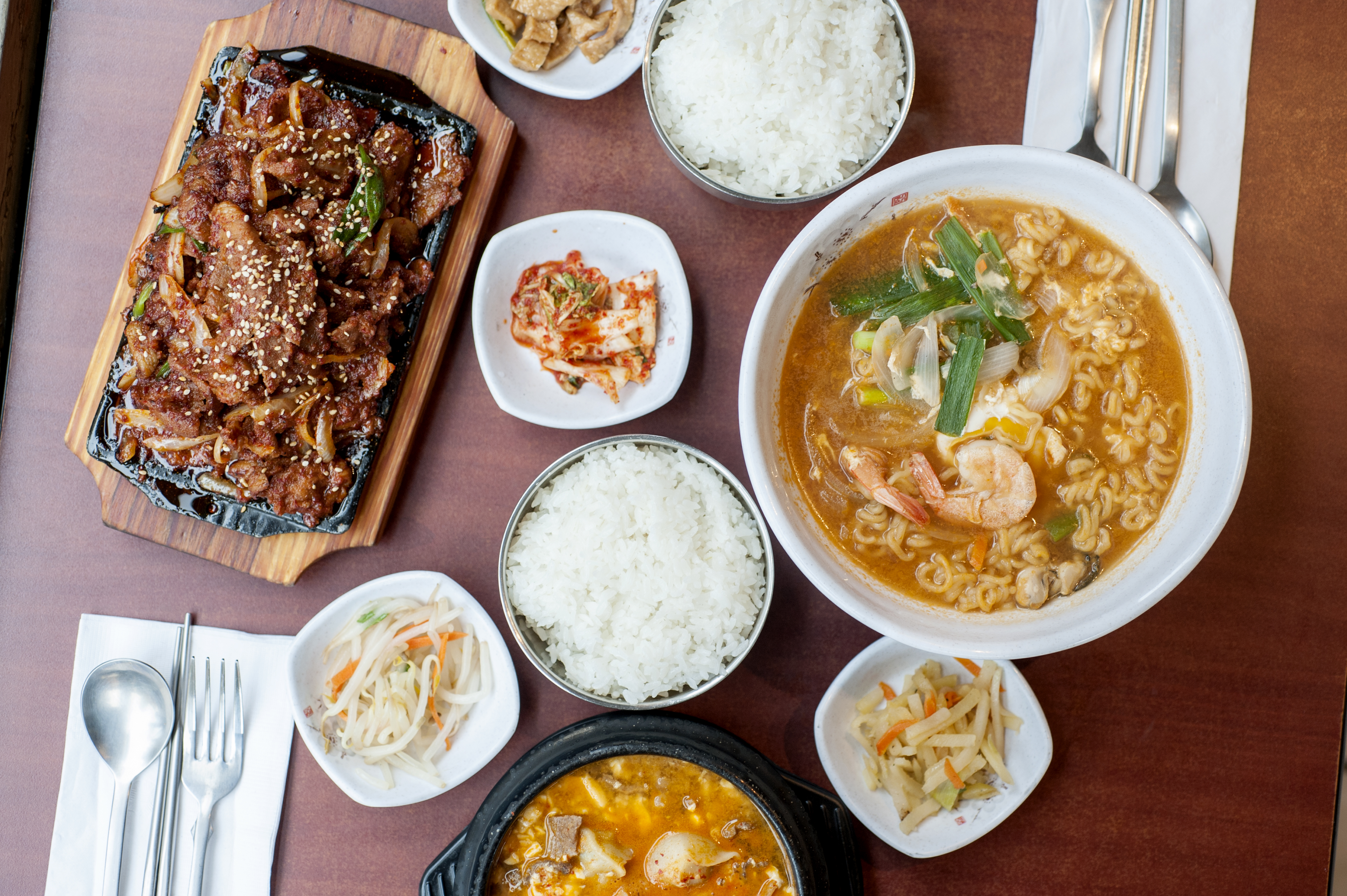 An assortment of white bowls holding various Korean dishes from an aerial view; they’re all placed on a dark red table. 