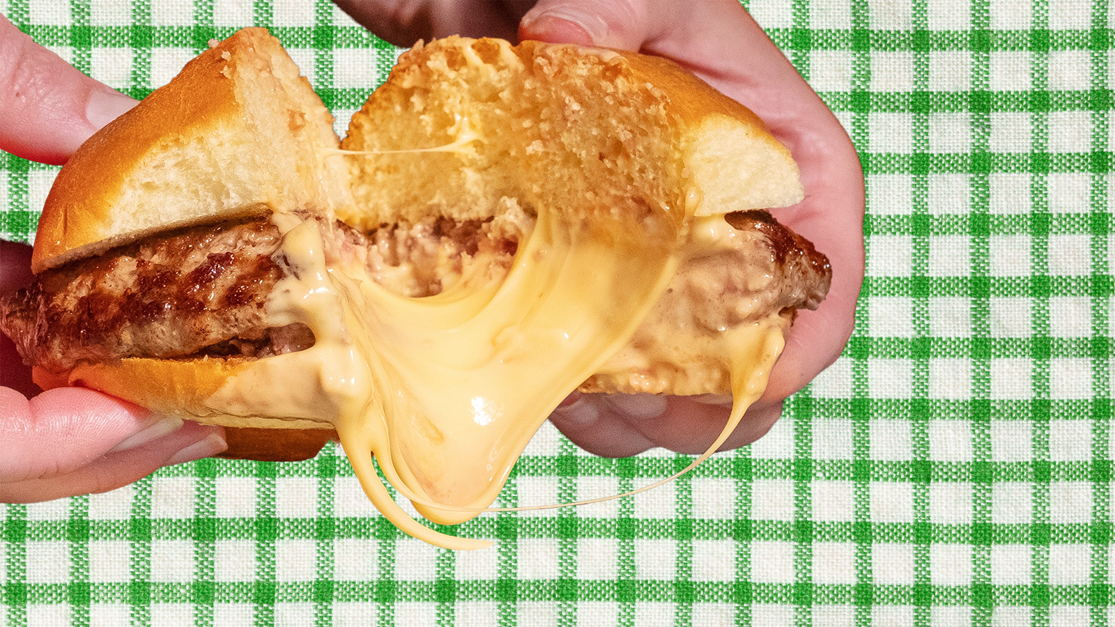 Photo illustration of a thick Juicy Lucy burger (dripping with molten American cheese in its center) over a green gingham background.