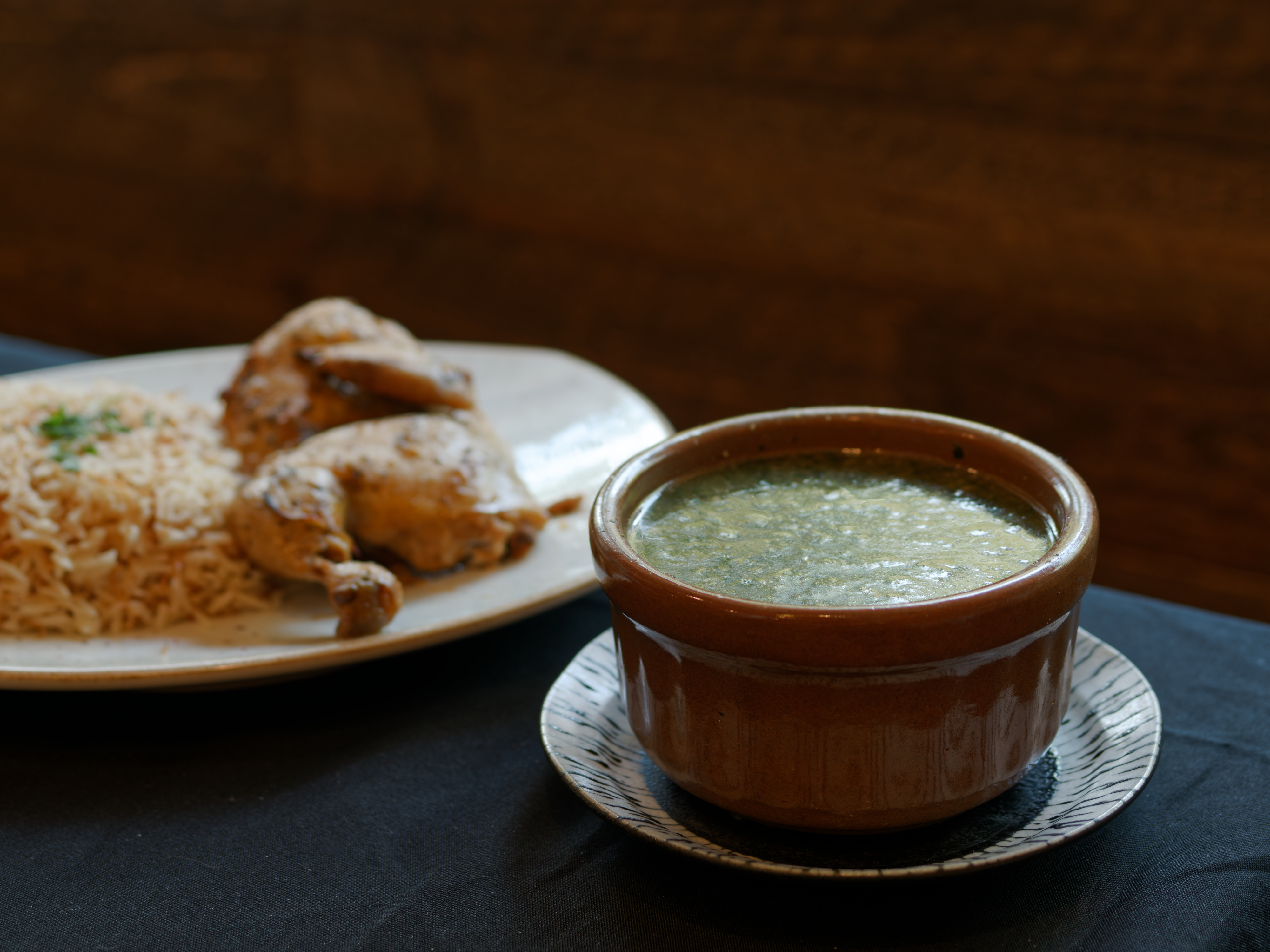 A bowl of molokhia in a brown bowl with a plate of a half of roasted chicken and rice in the background.