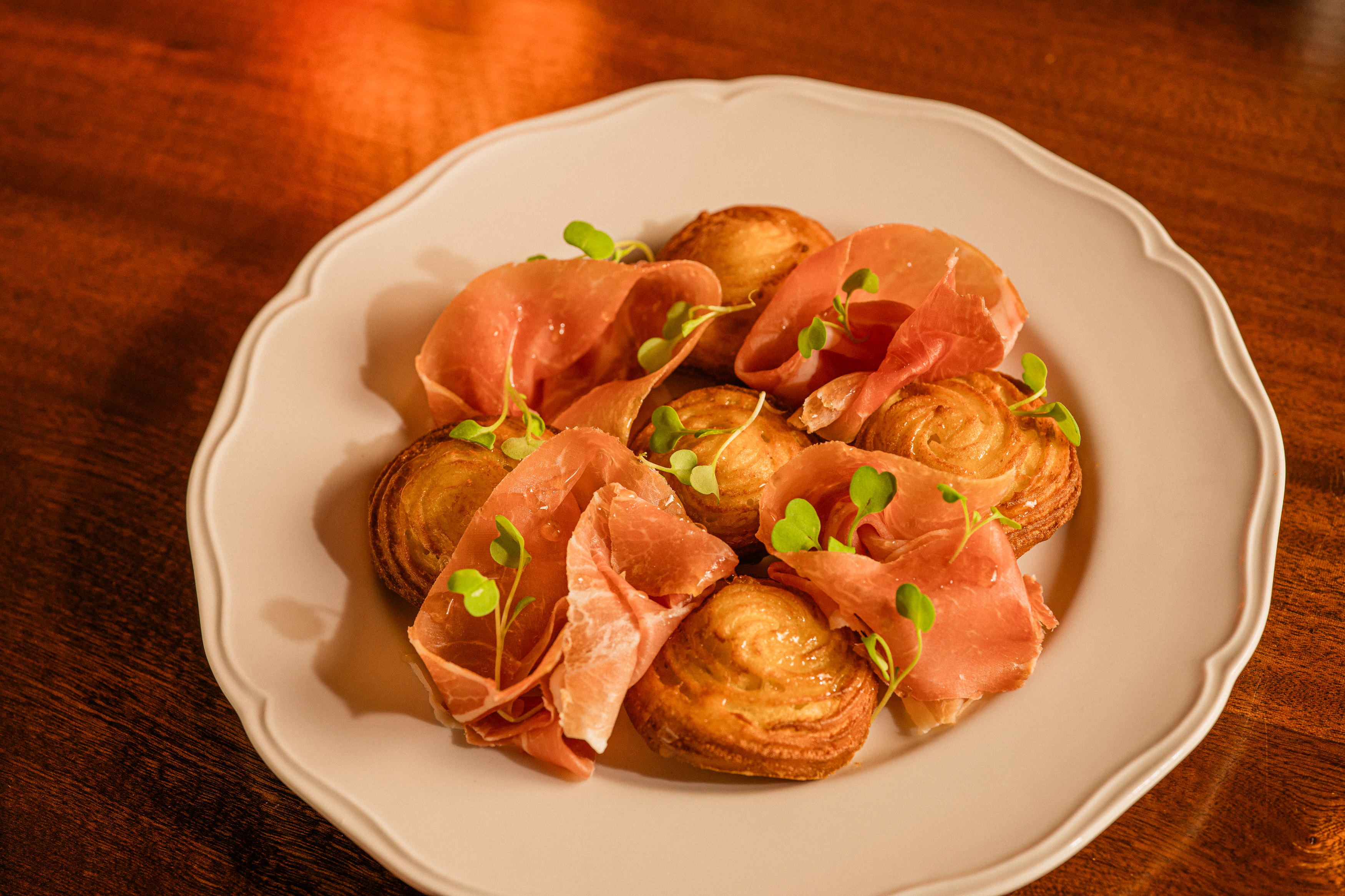 Golden doughnut puffs and shavings of prosciutto are arranged on a white plate with honey drizzled over top.