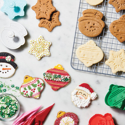 Christmas Pop Out Cookie Cutters with roll out cookies. Gingerbread and vanilla roll-out cookies on cooling rack without decorations in snowman, ornament, and snowflake. On counter top, completely decorated Santa, snowman, snowflake, and ornament cookies.